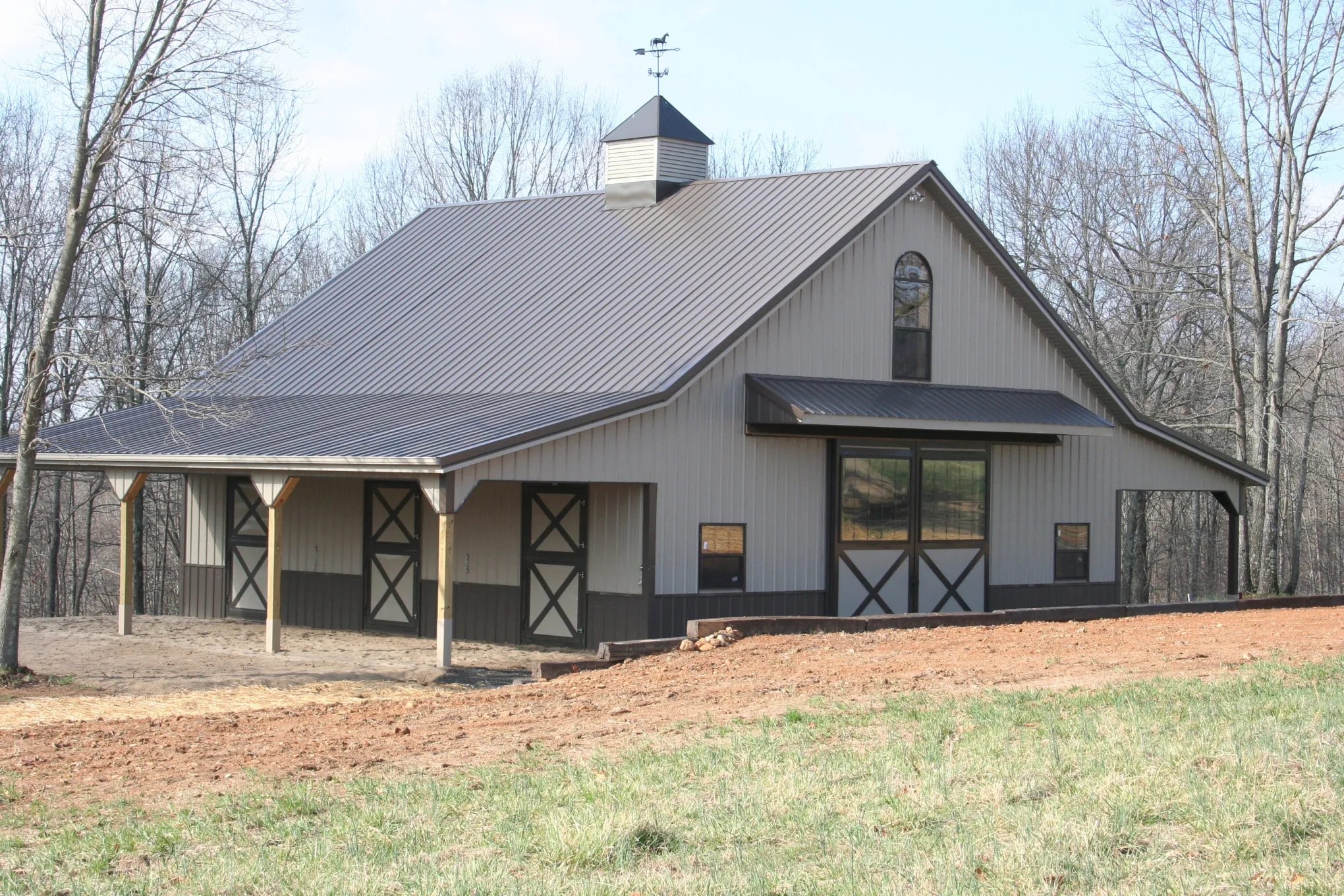 Tan post-frame horse barn with brown wainscot and a bronze metal roof, a white cupola with a horse weathervane, an arched gable window, and X-braced stall doors under a side overhang