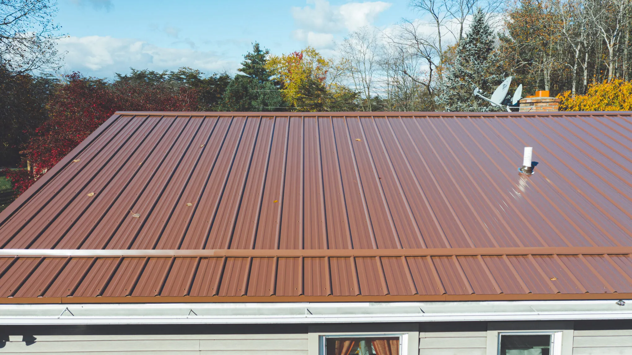 A close aerial view of a newly installed brown metal roof on a single-storey home with pale grey siding, autumn trees behind it