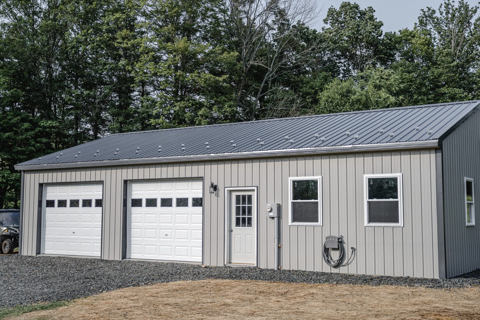 Post-frame garage with tan siding, a charcoal standing-seam metal roof, and two white overhead doors beside a white entry door