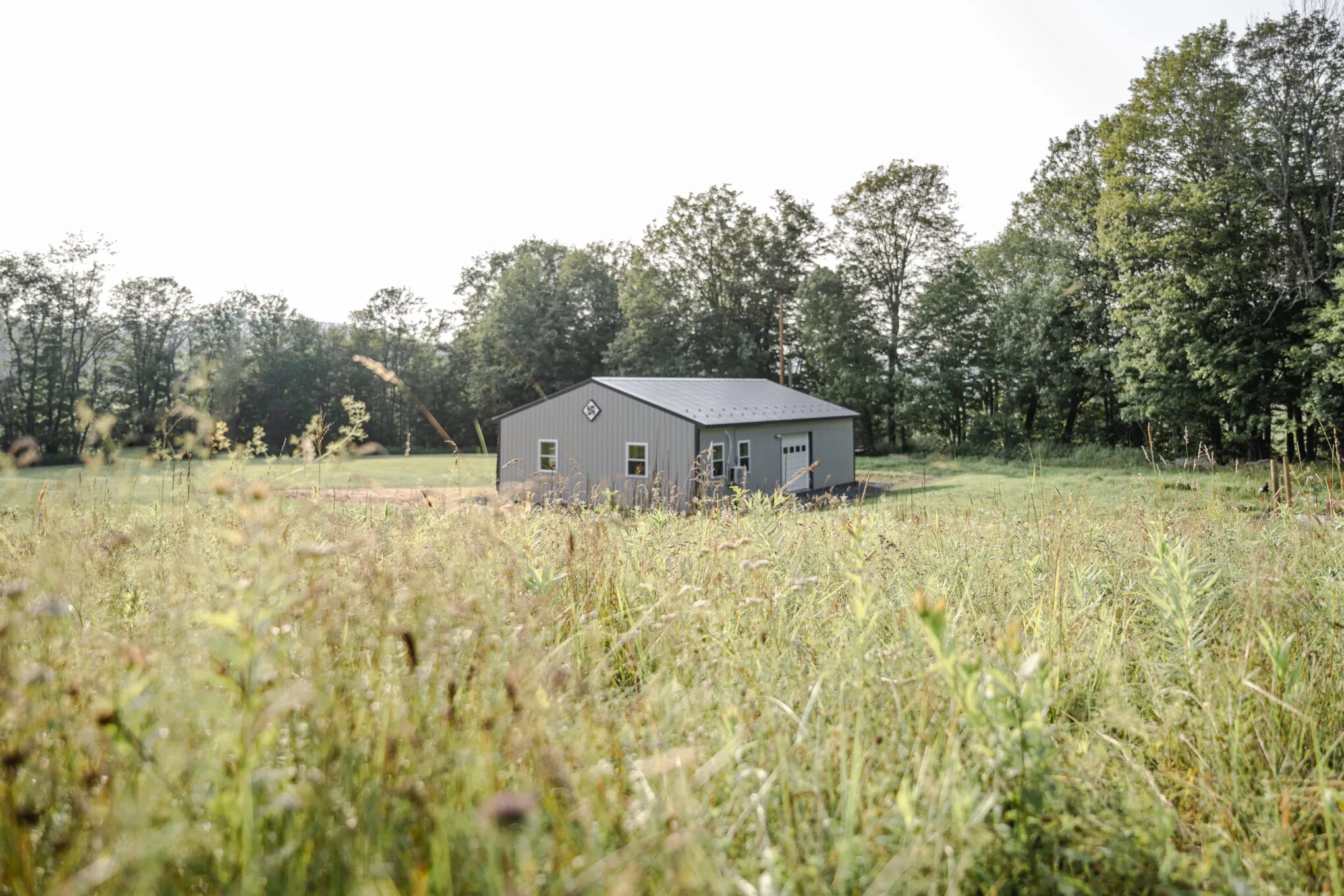 A small gray post-frame storage building with a charcoal roof and a barn quilt on the gable, seen across a wildflower meadow