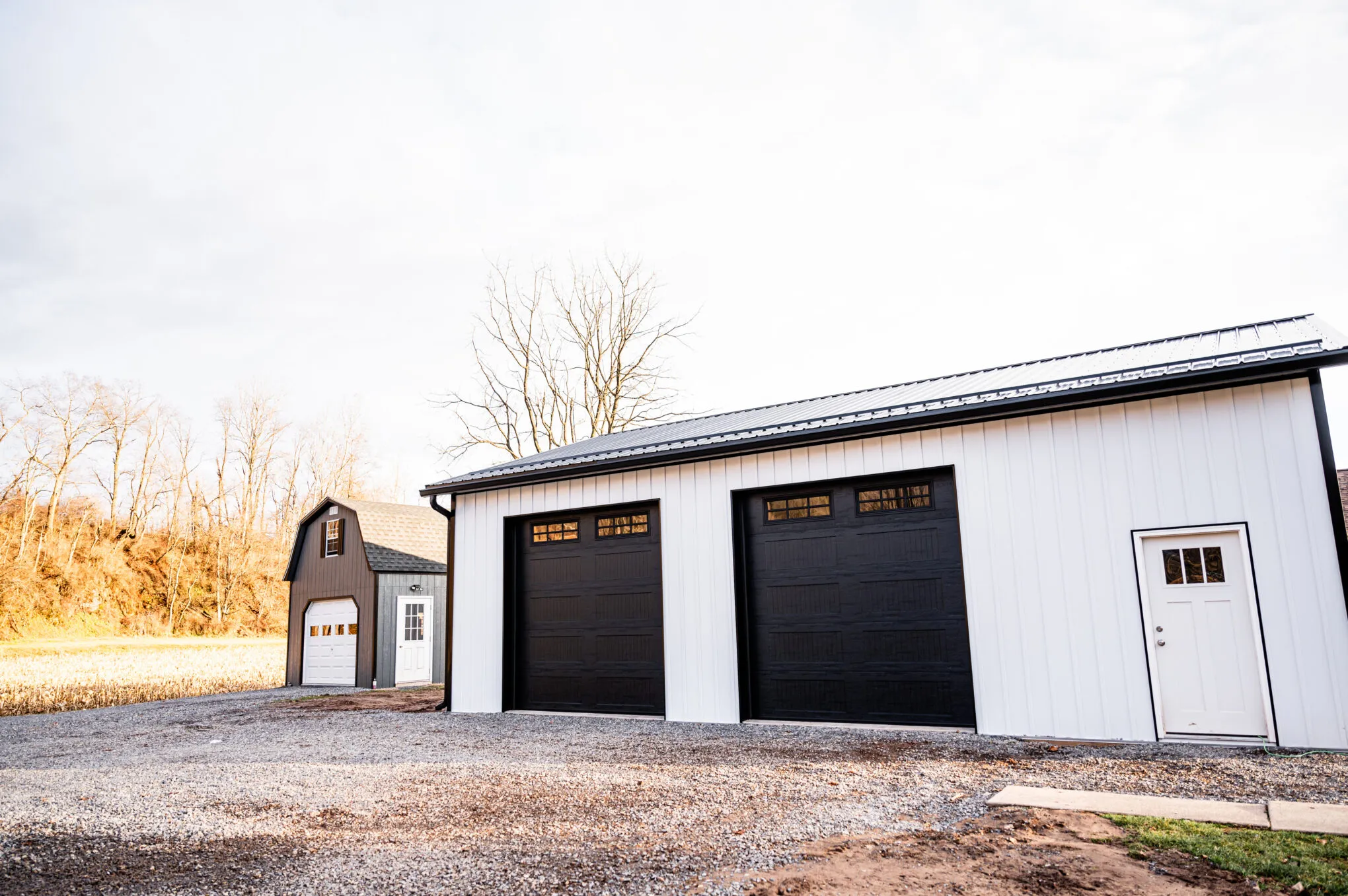 White post-frame garage with black wood-grain overhead doors and a white entry door, on a gravel drive with a barn behind