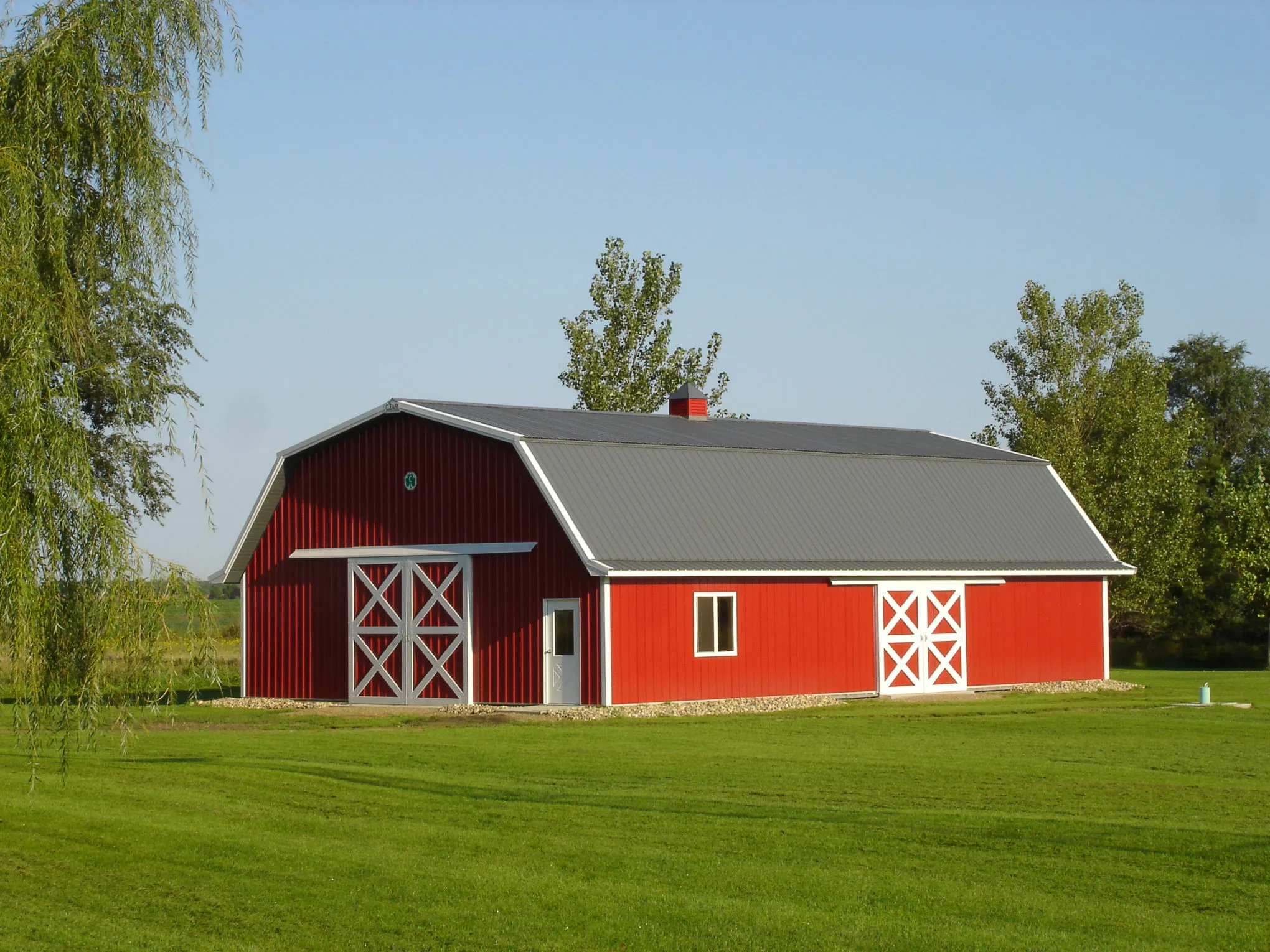Red gambrel-roof barn with a charcoal metal roof, white trim, and two sets of white cross-buck sliding doors, standing on a mown lawn