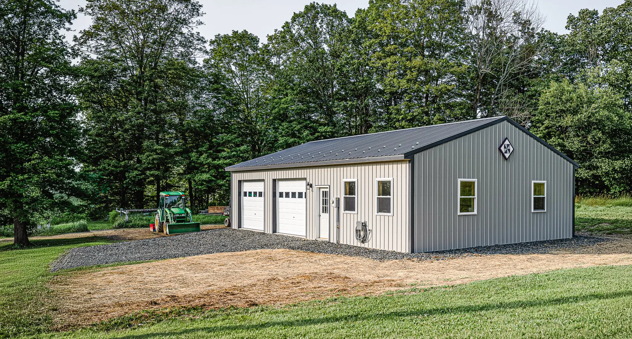 A gray post-frame garage with two white overhead doors and a dark metal roof, with a tractor parked beside it on a wooded Pennsylvania property